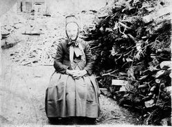 Alte Frau in Tracht neben Brennholz, wohl um 1900